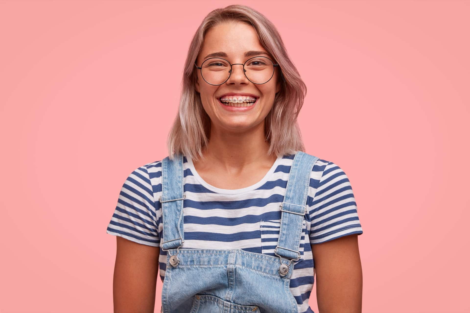 positive glad smiling female teenager with braces on teeth, wears stylish denim overalls and striped sailor t shirt, has broad smile, isolated over pink background. people, youth and positiveness