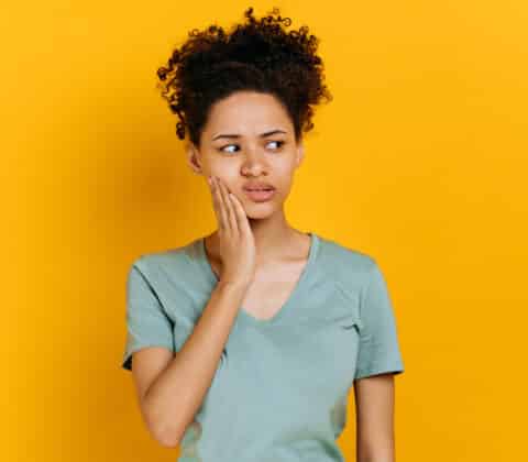 sad unhealthy african american woman touching her cheek with hand with painful expression because of toothache or dental illness on teeth, standing on orange isolated background. toothache concept